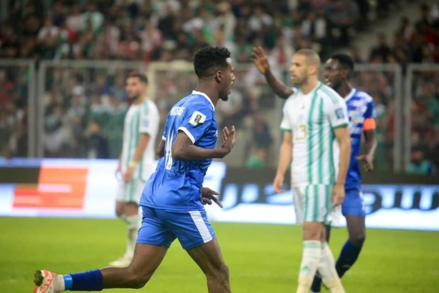 Ein Fußballer in einem blauen und weißen Trikot feiert ein Tor, umgeben von Teamkollegen in weißen und grünen Trikots und einer jubelnden Menge im Hintergrund.