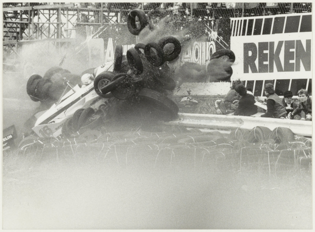 Schwarz-weiß-Foto eines Rennwagens, der in einen Zaun gekracht ist und von Reifen umgeben ist, mit Menschen zu seiner Rechten und einer Tafel mit Text im Hintergrund.