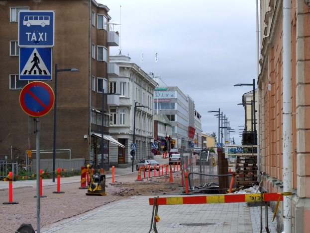 Stadtstraße mit Gebäuden, Straßeninfrastruktur, Fahrzeugen, einer Baustelle mit Verkehrsschildern, Bäumen und einem bewölkten Himmel.
