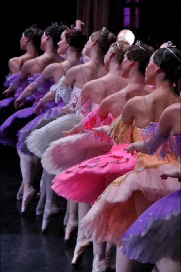 Balletttänzerinnen in rosafarbenen Tutus sitzen auf einer Bühne, beleuchtet von einem Licht im Hintergrund, bewegen sich in synchronisierten Posen.