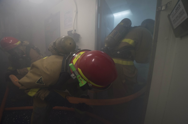 Feuerwehrleute in Schutzausrüstung betreten einen raucherfüllten Raum mit Rohren und einer Lampe im Hintergrund.