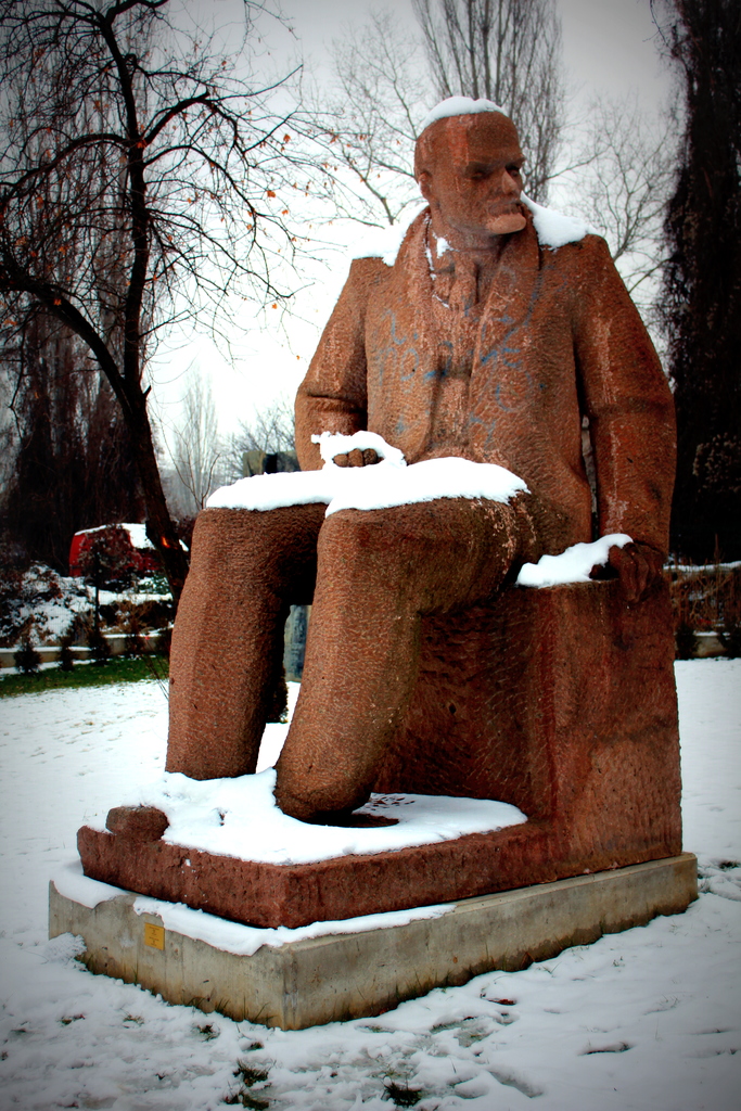 Eine Skulptur einer sitzenden Person umgeben von Schnee mit B├Ąumen im Hintergrund.