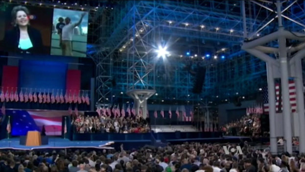 Große Menschenmenge vor einer Bühne mit einem Rednerpult, Mikrofon, Flaggen, einem Bildschirm und Beleuchtungsausrüstung, was auf eine Präsidentschaftsdebatte hindeutet.