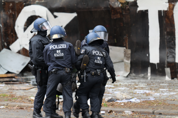 Polizisten in schwarzen Uniformen und blauen Helmen stehen vor einem Graffiti-bedeckten Gebäude während einer Unruhen, mit verstreuten Papieren und Gegenständen auf dem Boden.