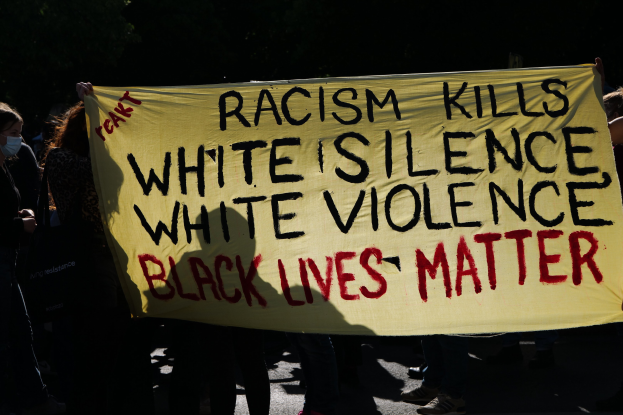 Eine Gruppe maskierter Personen mit einem Banner "Rassismus tötet Weißes Schweigen, weiße Gewalt, Black Lives Matter" auf einer dunklen Straße.