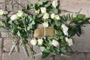 Wei├čer Rosenkranz mit grünen Bl├Ąttern und zwei goldenen, beschrifteten Gegenst├Ąnden auf dem Boden vor einem Berliner Mahnmal.