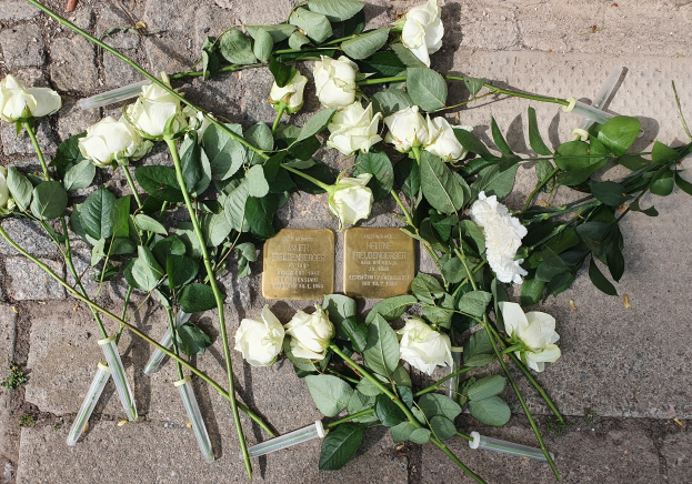Wei├čer Rosenkranz mit grünen Bl├Ąttern und zwei goldenen, beschrifteten Gegenst├Ąnden auf dem Boden vor einem Berliner Mahnmal.