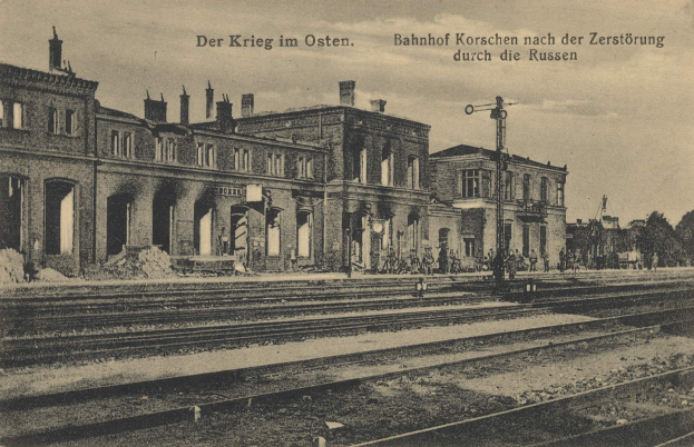 Schwarzes und weißes Foto des Bahnhofs Korschen in Ostern, Deutschland, das Schienen, Bahnhofsgebäude, Menschen, Bäume und einen Himmelhintergrund mit dem Text "Bahnhof Korschen in Ostern, Deutschland" oben und unten zeigt.