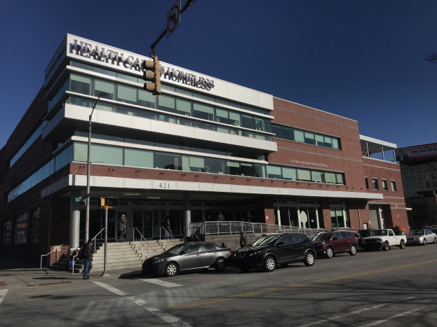 Großes Gebäude, das als New York City Health Care Homeless Center etikettiert ist, mit geparkten Autos, Fußgängern, Straßenmöblierung und Bäumen unter einem klaren Himmel.