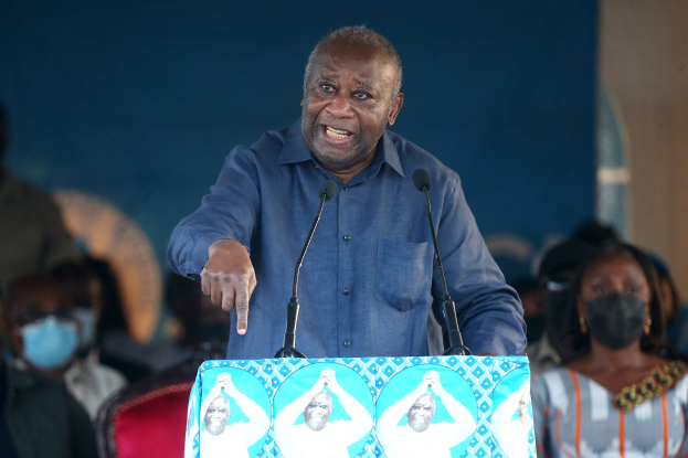 Ein Mann in einem blauen Hemd spricht in zwei Mikrofone an einem Rednerpult und wendet sich an eine Menge, einige tragen Masken, mit einer Wand im Hintergrund.