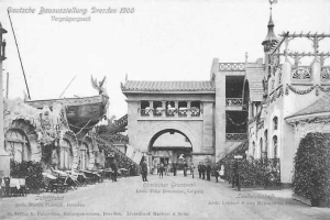 Ein Schwarz-Weiß-Foto aus dem Jahr 1900, das Menschen zeigt, die eine Straße in Dresden, Deutschland entlanggehen, gesäumt von Gebäuden, Pflanzen und Stühlen, mit Text oben und unten auf dem Bild.