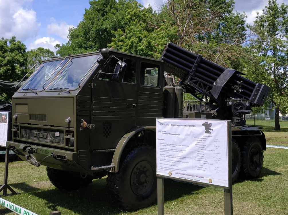Ein Militärlastwagen mit einer Rakete darauf steht auf einer grasbewachsenen Fläche umgeben von Bäumen unter einem klaren blauen Himmel, mit einer Texttafel davor, die wahrscheinlich Teil einer russischen Militärausstellung ist.