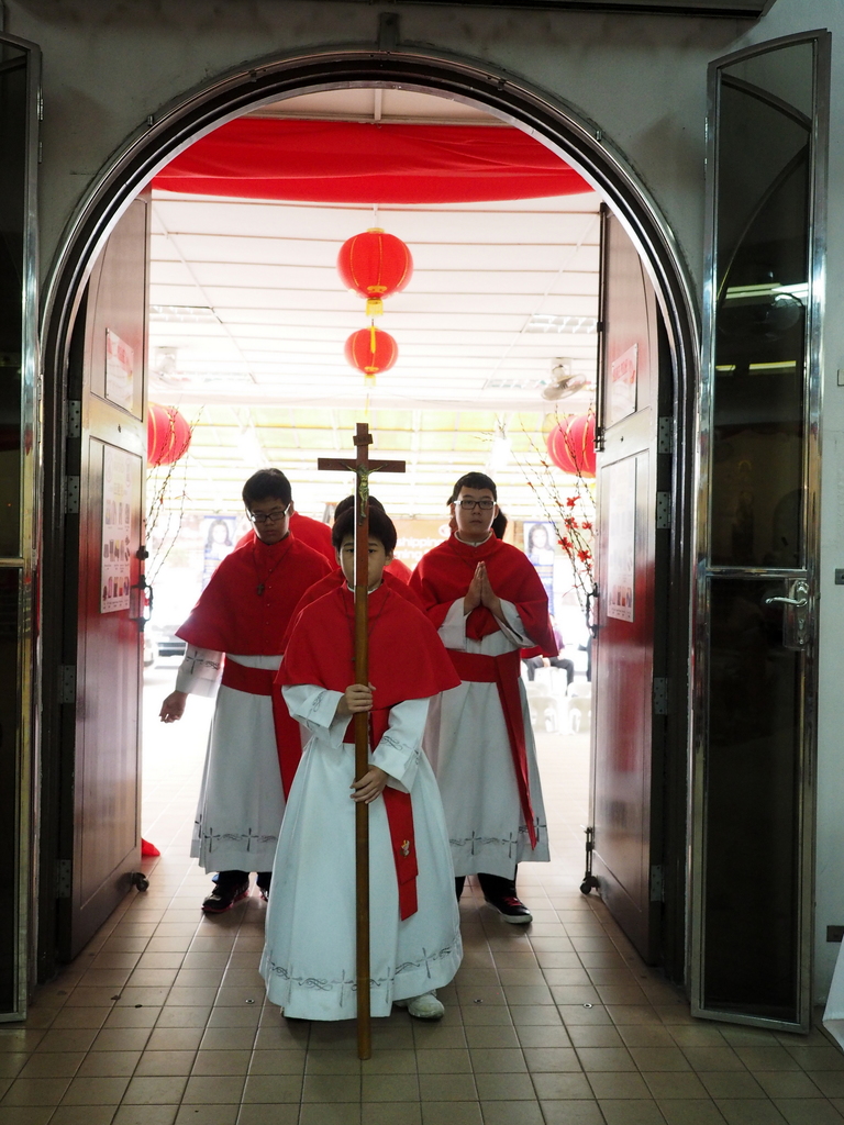 Eine Gruppe von Menschen, die mit einem Kreuz in der Mitte einer Kathedrale gehen, mit Türen links und rechts und Blumen und dekorativen Elementen im Hintergrund.