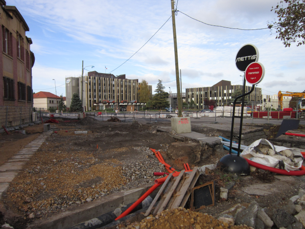 Baustelle mit verstreuten Materialien (Steine, Holz, Rohre), Schilder an Pfählen, ein Zaun, Fahrzeuge, Bäume, Gebäude mit Fenstern, Laternenmasten und eine bewölkte Himmelslandschaft im Hintergrund.
