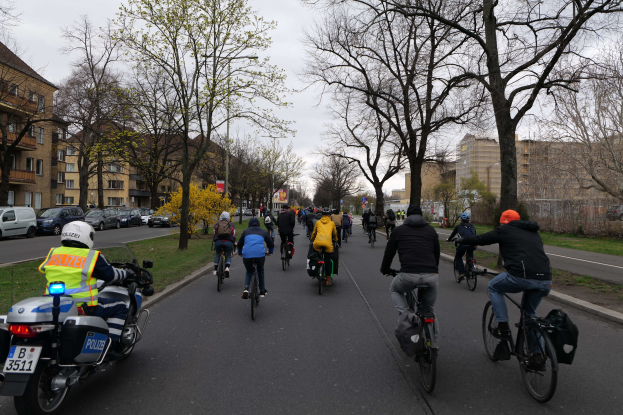 Eine Gruppe von Menschen, die auf Fahrrädern eine von Bäumen und Gebäuden gesäumte Straße entlangfährt, mit einem Polizeibeamten auf einem Motorrad links daneben.