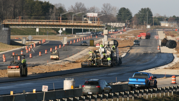 Baustelle mit Fahrzeugen auf der Straße, Verkehrszeichen, Schilder, Pfosten, eine Brücke, Bäume und einen klaren blauen Himmel.