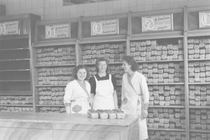 Drei Frauen in Schürzen lächeln vor einem Bäckertisch mit Lebensmitteln, Gestellen und Wandtafeln im Hintergrund, dargestellt in Schwarz-Weiß.